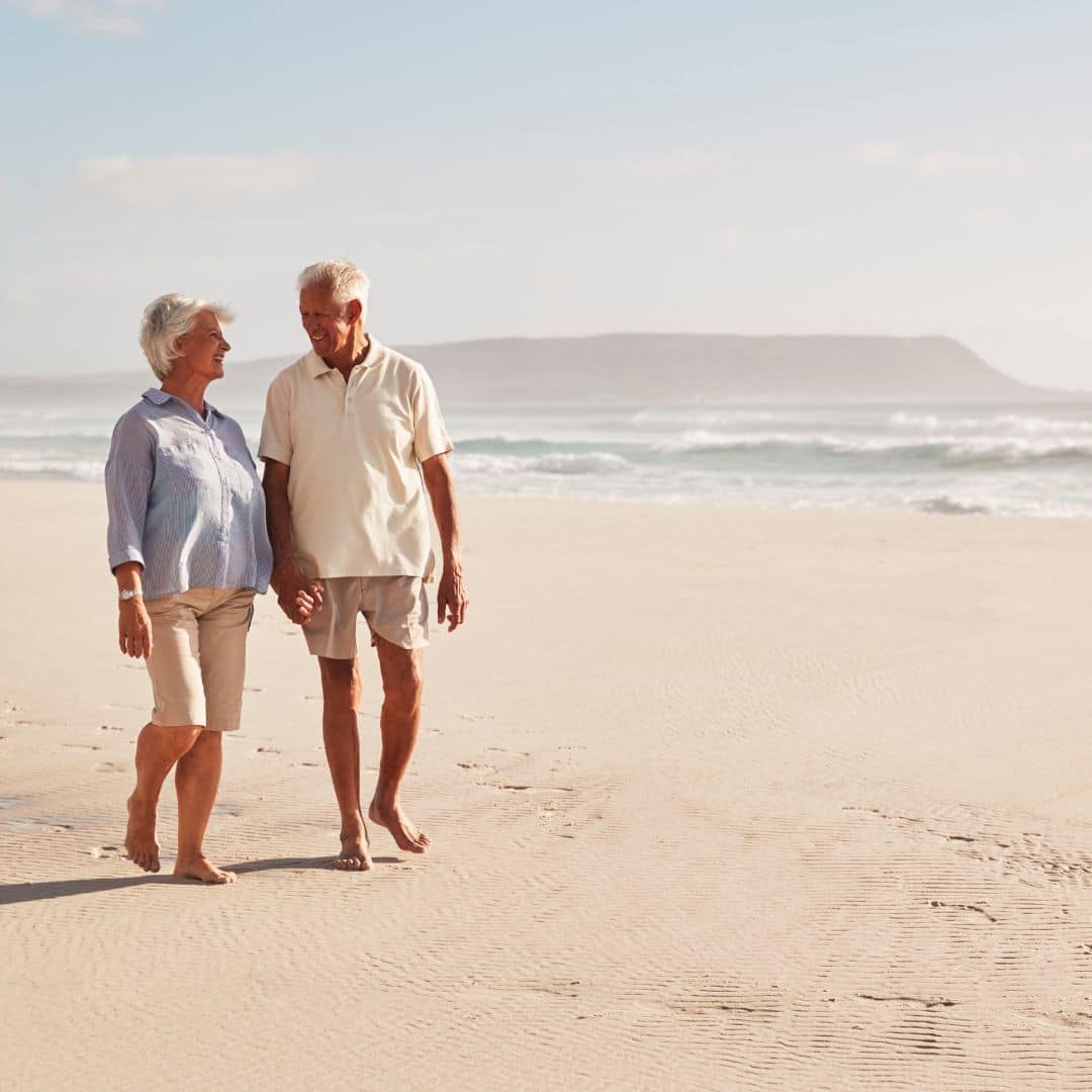 An elderly couple enjoying a walk along the beach with the surf rising in the background.