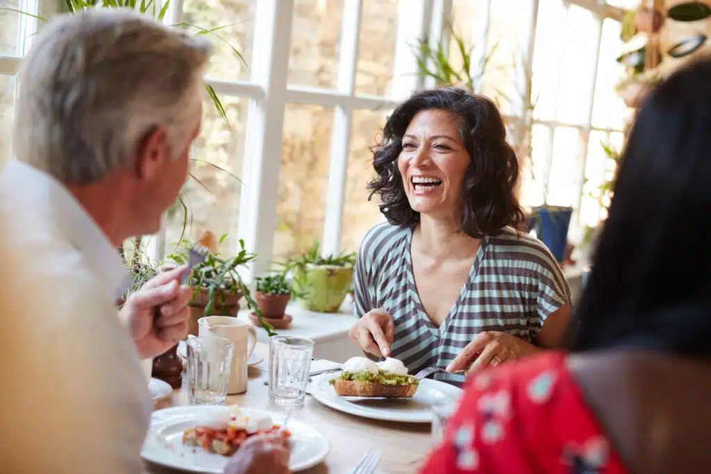 A woman laughs joyfully while enjoying a meal with friends at a bright, plant-filled café. Natural light streams through large windows behind her, illuminating plates of food and drinks on the table as they share a cheerful moment.