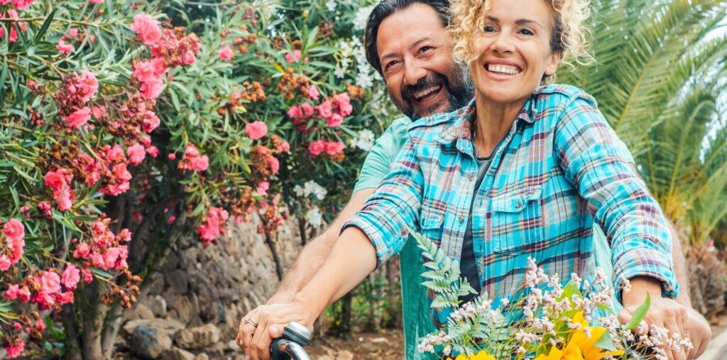 A joyful couple rides a bicycle together along a garden path, with the woman in front smiling brightly and a basket of fresh flowers attached to the handlebars. Colorful pink blooms and palm fronds surround them, creating a vibrant, tropical atmosphere.