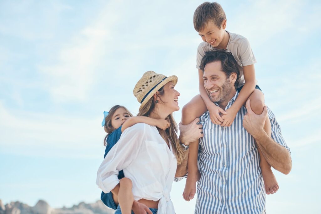 A cheerful family enjoys a sunny day outdoors, with the mother carrying a young girl on her back and the father holding a boy on his shoulders. They are all smiling and looking at each other warmly, with a clear blue sky and rocky landscape in the background.