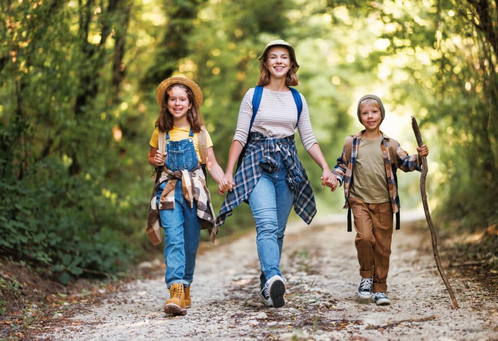 A smiling woman walks hand in hand with two young children down a forest trail, all dressed in casual outdoor clothes with flannel shirts and hats. The girl wears denim overalls and a straw hat, while the boy holds a walking stick, surrounded by lush green trees and dappled sunlight.