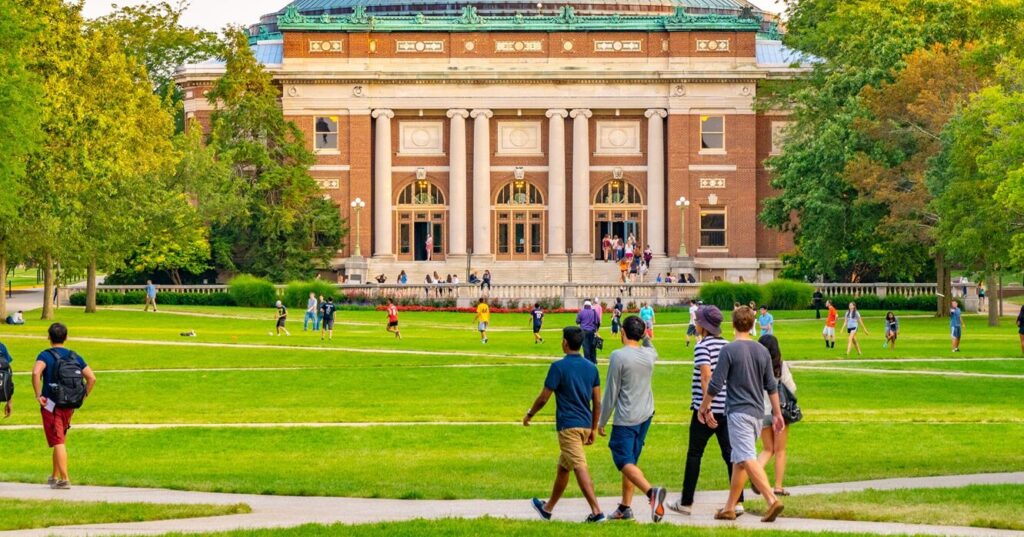 A large group of students walks and lounges on a lush green lawn in front of a grand, domed brick building with tall columns and arched windows. The scene captures a lively college campus atmosphere on a sunny day, surrounded by trees and academic architecture.
