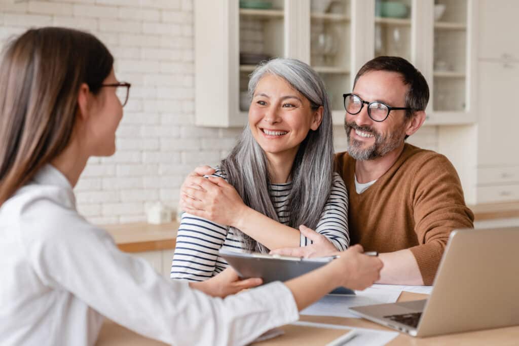 A smiling couple sits closely together at a table with a retirement advisor, looking optimistic and engaged. The woman warmly embraces her partner as they review documents, suggesting trust and satisfaction. A laptop and papers in front of them reflect the planning process for their financial future.
