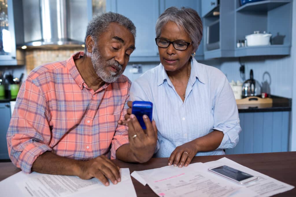 An older couple sits at a kitchen table reviewing bills and paperwork, looking at a blue calculator together. They appear focused as they work through financial documents spread across the table.