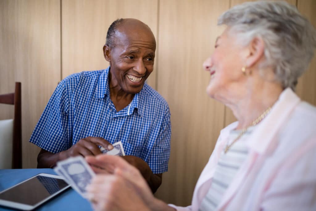 An older man and woman share a laugh while playing cards at a table. The man, wearing a blue checkered shirt, smiles warmly as he holds his cards, while the woman, slightly out of focus, looks back at him with joy.