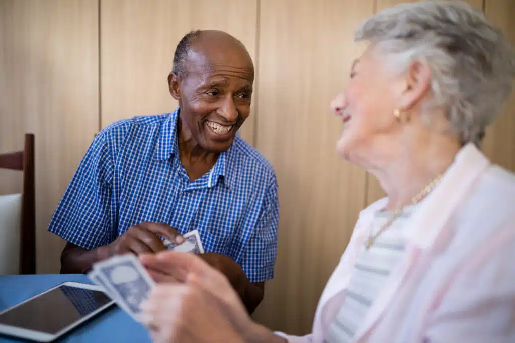 An older man and woman share a laugh while playing cards at a table. The man, wearing a blue checkered shirt, smiles warmly as he holds his cards, while the woman, slightly out of focus, looks back at him with joy.
