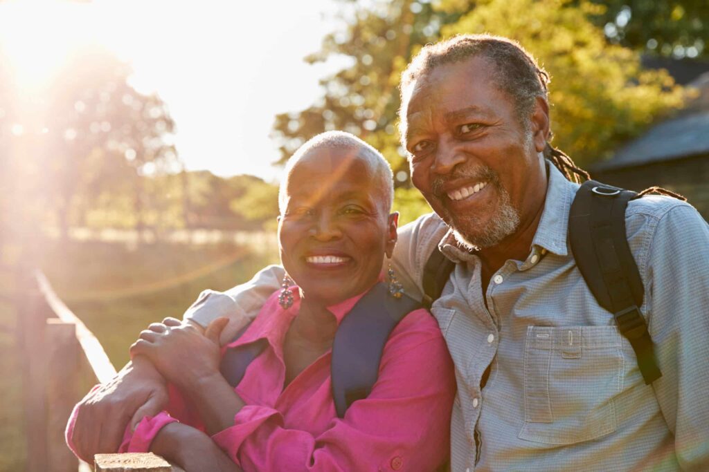 An older couple stands outdoors in the sunlight, smiling warmly at the camera. The woman wears a bright pink shirt and leans on the man's arm, while he has his arm around her shoulder with a backpack on.