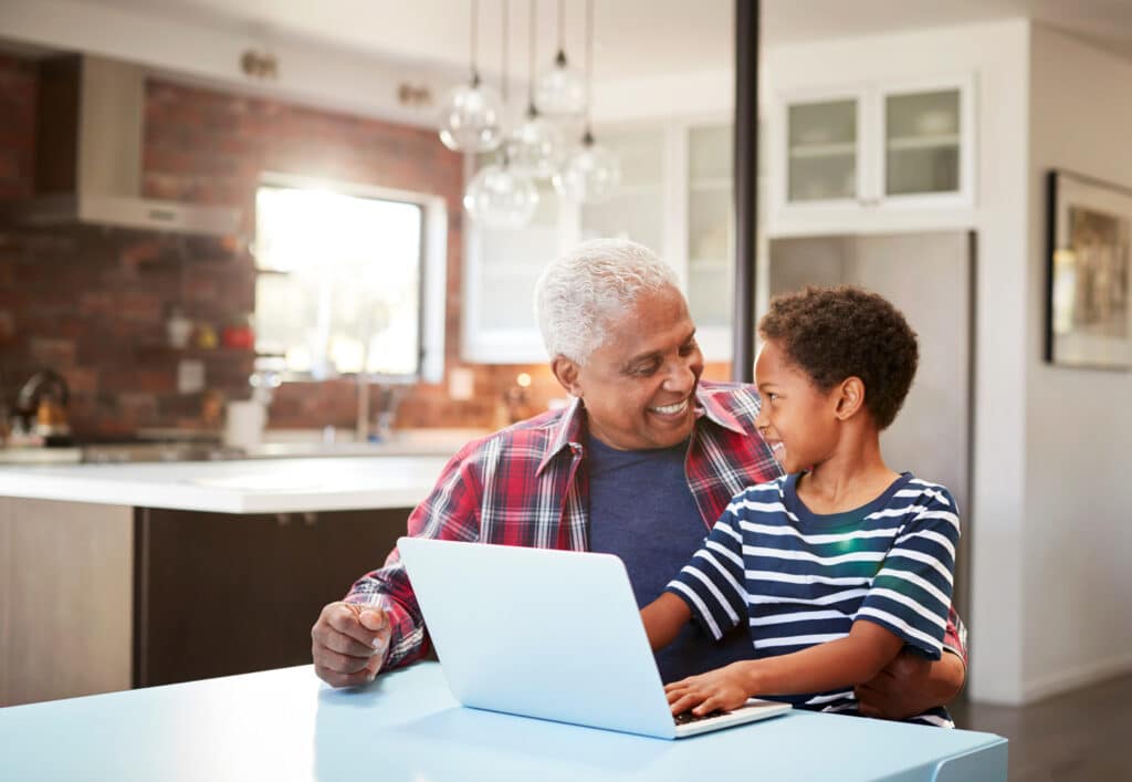 An older man and his grandson sit together at a table with a laptop, smiling warmly at each other. The boy rests his hands on the keyboard while the man has his arm around him in a modern kitchen setting.