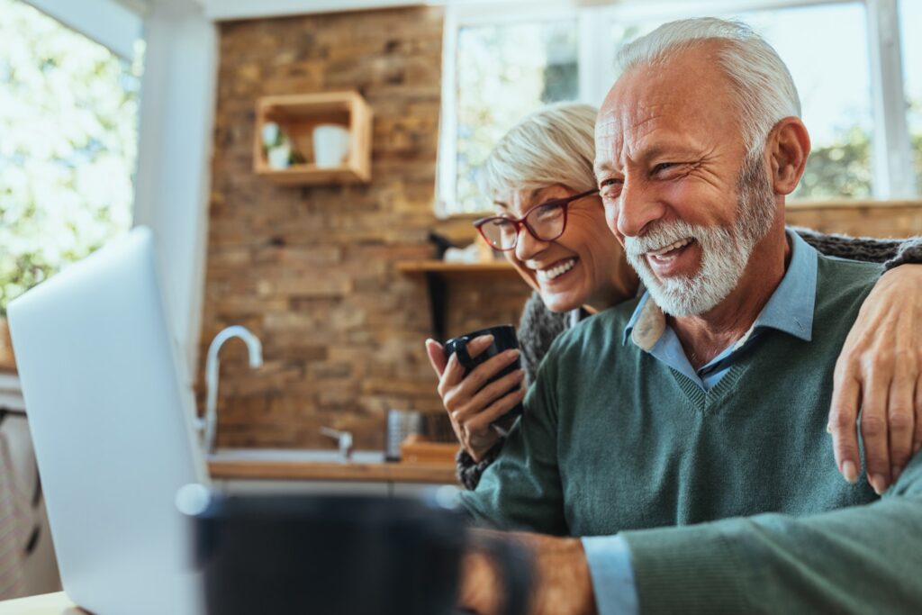An older couple sits together in a cozy kitchen, smiling and laughing as they look at a laptop screen. The woman holds a coffee mug and leans close with her arm around the man.
