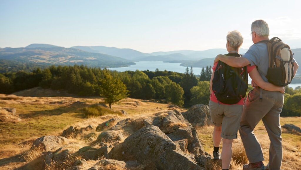 An older couple with backpacks stands on a rocky hilltop, arms around each other, admiring a wide view of a lake surrounded by forests and mountains. The warm sunlight highlights the scenic landscape, suggesting a peaceful hiking moment.