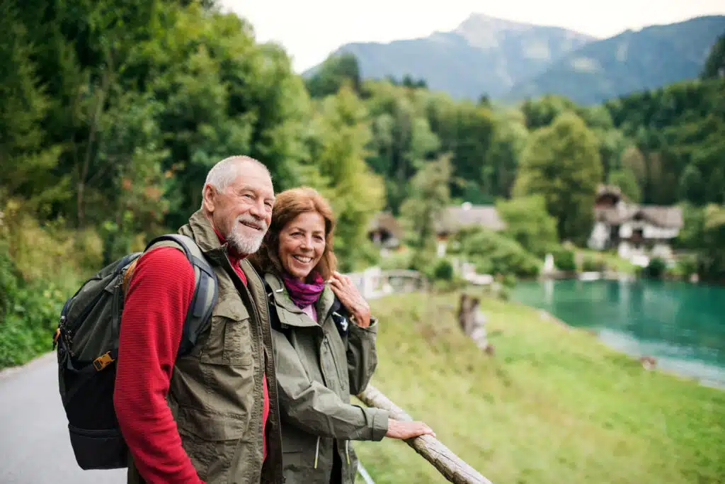 An older couple stands by a wooden railing, smiling as they enjoy a scenic view of a turquoise lake surrounded by trees and mountains. They are dressed in outdoor gear with backpacks and jackets, suggesting a hike or nature walk.