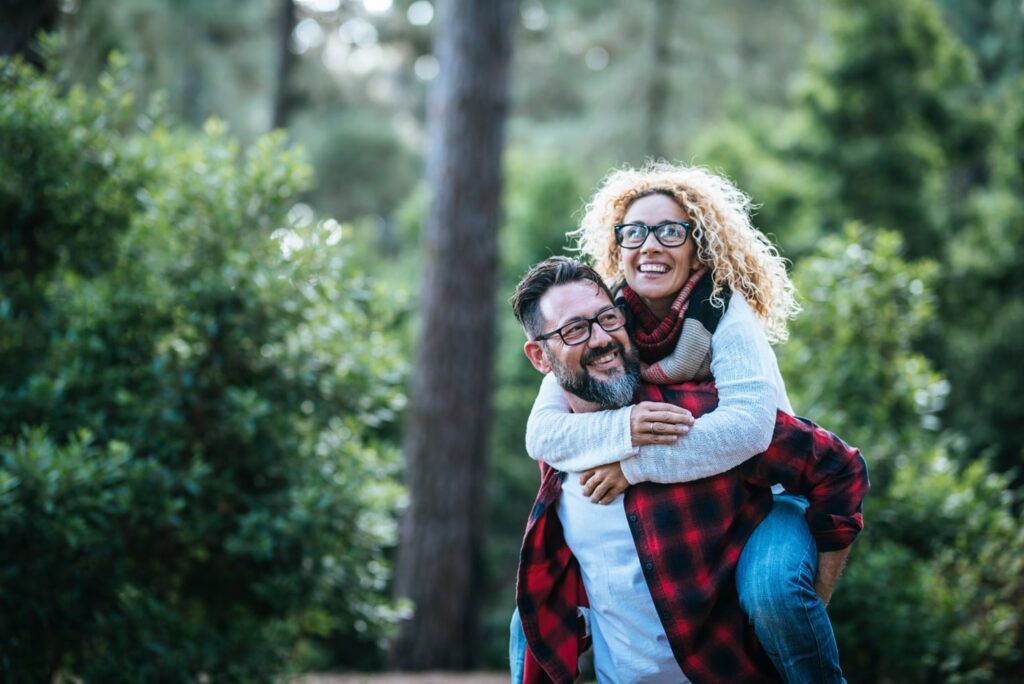 A joyful couple shares a playful moment in a forested area, with the woman riding piggyback on the man’s back. Both are smiling and wearing glasses and cozy layers, surrounded by tall trees and lush greenery.