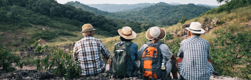 Four older adults wearing hats and backpacks sit side by side on a rocky hilltop, looking out over a lush green valley and distant mountains. They appear relaxed, enjoying the scenic view together.