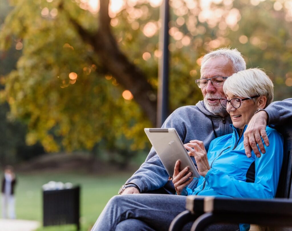 An older couple sits together on a park bench, smiling as they look at a tablet. The man has his arm around the woman while autumn trees glow softly in the background.