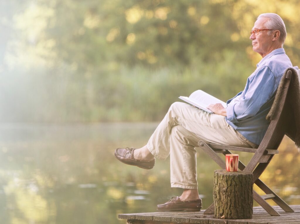 An older man sits in a wooden chair by a peaceful lake, reading a book with a relaxed expression. A red coffee cup rests on a tree stump beside him as soft sunlight filters through the trees in the background.