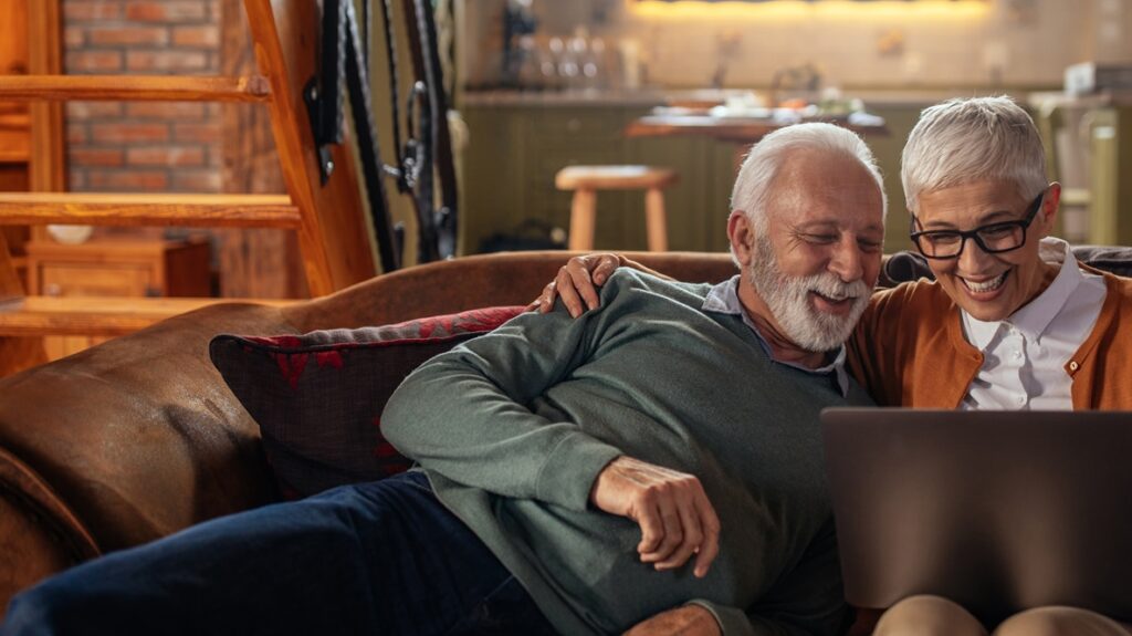 An older couple sits together on a cozy couch, laughing as they look at a laptop screen. The warm lighting and homey background create a relaxed and cheerful atmosphere.