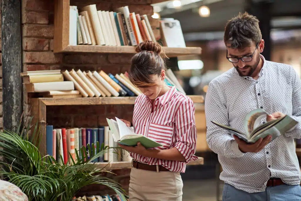 A woman and man stand side by side in a cozy bookstore, each absorbed in their own book. Surrounded by wooden bookshelves filled with colorful spines, they appear relaxed and focused under soft lighting.