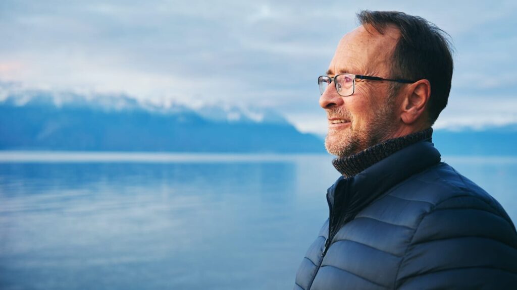 An older man wearing glasses and a puffer jacket smiles as he looks out over a calm lake. Snow-capped mountains and a cloudy sky create a peaceful backdrop.