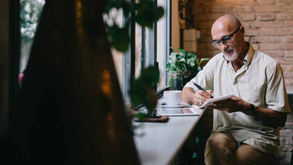 An older man with glasses sits by a café window, writing in a notebook while a cup of coffee rests on the counter beside him. Soft light and indoor plants create a calm, focused atmosphere.