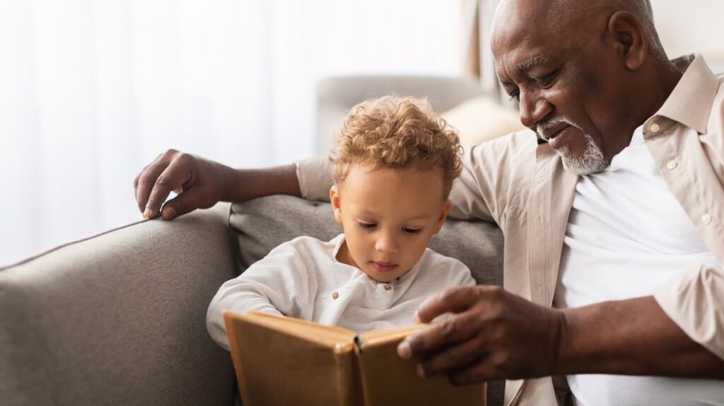 An older man sits on a couch with his grandson, reading a book together. The child looks focused on the pages while the man smiles warmly, holding the book open.