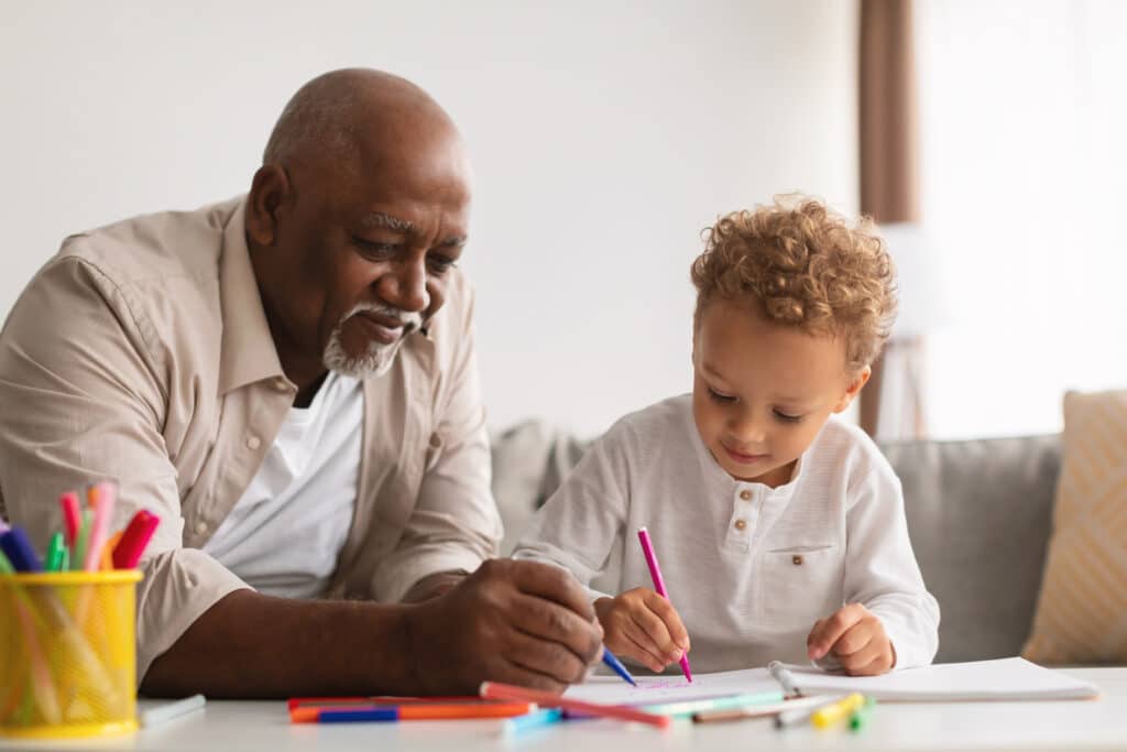 An older man sits beside a his grandson at a table, both focused on drawing with colorful markers. The man smiles gently while the child holds a pink marker, surrounded by scattered pens and papers.
