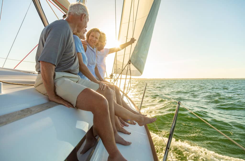 A group of older adults sit along the edge of a sailboat, smiling and enjoying the breeze as the sun sets over the water. Their bare feet dangle above the waves, with sails catching the light behind them.
