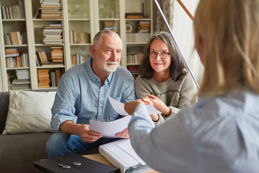 An older couple is sitting together at a table, meeting with an advisor. The man is holding paperwork while the woman sits closely beside him, smiling warmly. The retirement advisor, whose back is to the camera, is handing them documents. The setting feels comfortable and personal.