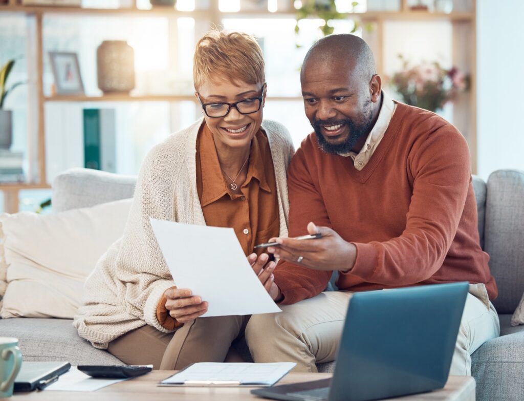 A smiling couple sits together on a couch, reviewing paperwork while pointing at details on the page. A laptop, calculator and documents are spread out on the coffee table in front of them.