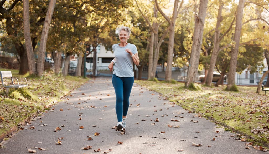 An older woman jogs along a tree-lined park path on a sunny day, smiling as she exercises. Fallen leaves scatter across the trail, adding to the autumn atmosphere.