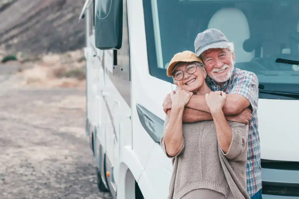 An older couple stands by their RV, smiling warmly at the camera while leaning close. They both wear casual clothes and caps, with a desert-like landscape in the background.