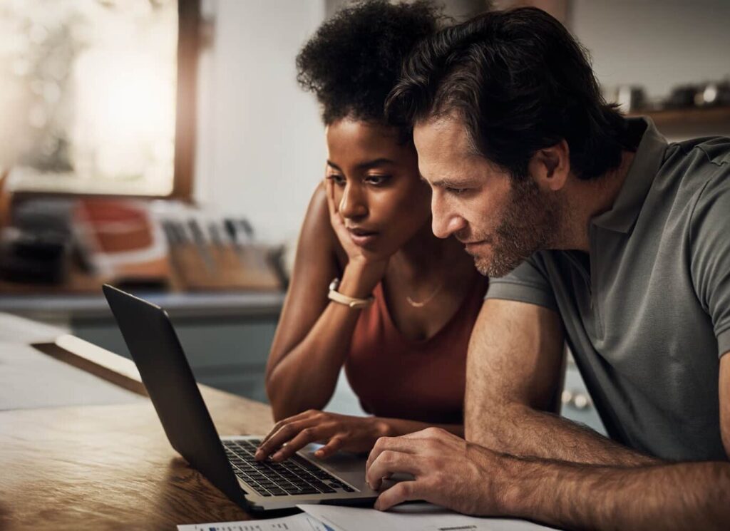 A couple leans in closely over a laptop at a wooden table, intently focused on the screen as they review something together. Soft natural light filters through a nearby window, creating a warm and thoughtful atmosphere.