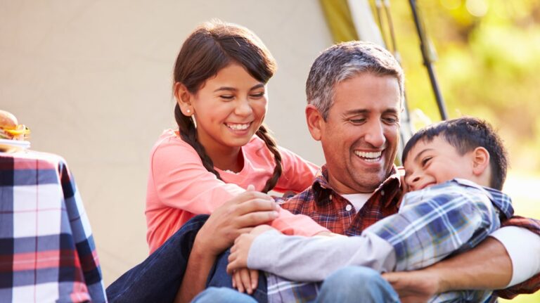 A father sits outdoors with his two children, laughing and playing together. The daughter, smiling brightly, leans on him while the son giggles in his arms. They look joyful and close, enjoying a playful family moment.