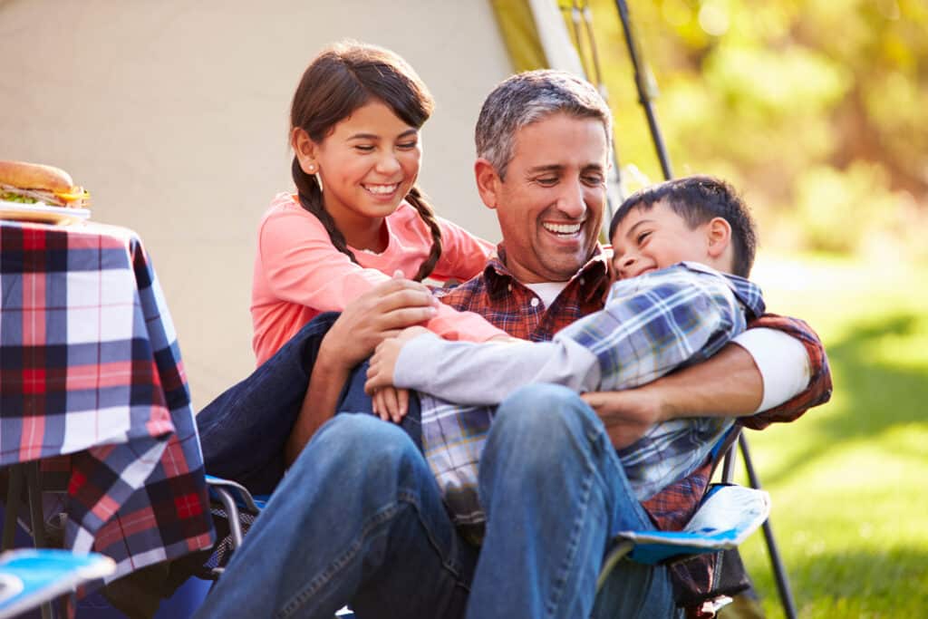 A father sits outdoors with his two children, laughing and playing together. The daughter, smiling brightly, leans on him while the son giggles in his arms. They look joyful and close, enjoying a playful family moment.