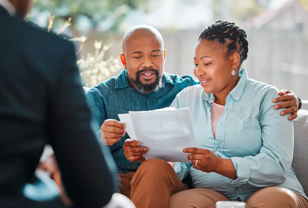 A couple sits closely together reviewing paperwork, smiling as they look at the documents in their hands. The man has his arm around the woman while a retirement advisor, slightly out of focus, faces them in the foreground.