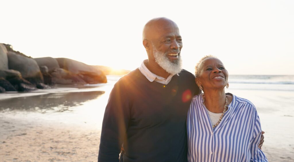 An older couple walks arm in arm along the beach at sunset, smiling as they look ahead. The man wears a dark sweater and the woman wears a blue and white striped shirt, with the sunlight glowing warmly behind them.