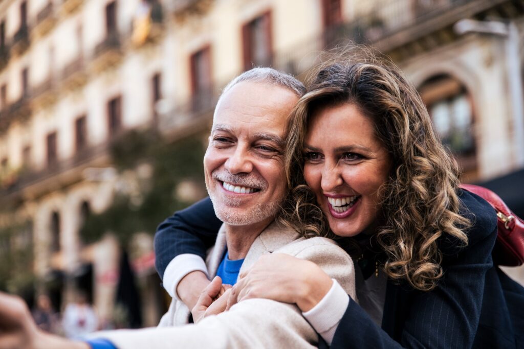 A smiling couple embraces closely while taking a selfie on a city street, with blurred European-style buildings in the background. The woman laughs joyfully over the man's shoulder, capturing a happy, candid moment together.