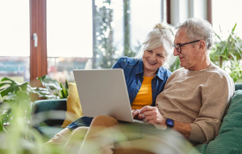A cheerful older couple sits together on a cozy couch, sharing a laptop. They appear relaxed and engaged, smiling as they browse or plan something online. Surrounded by greenery and natural light, the scene radiates comfort, contentment, and the ease of enjoying retirement life together at home.