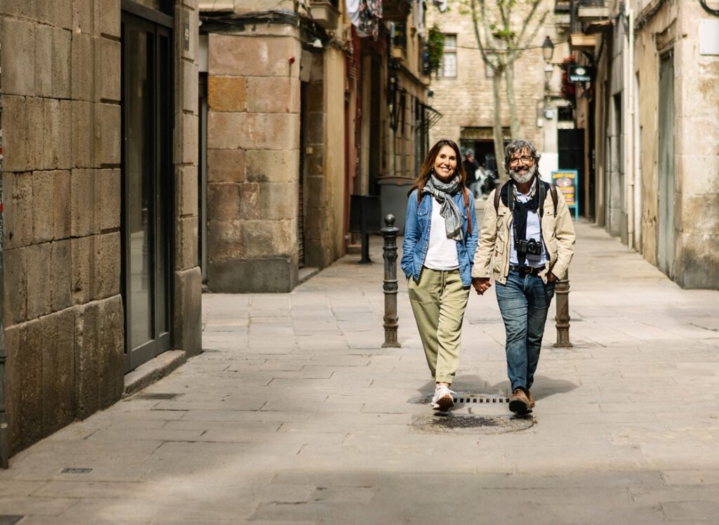 A smiling couple walks hand in hand down a quiet, sunlit European street lined with stone buildings. Both are dressed casually with backpacks, and the man has a camera hanging around his neck, suggesting they are exploring or traveling.