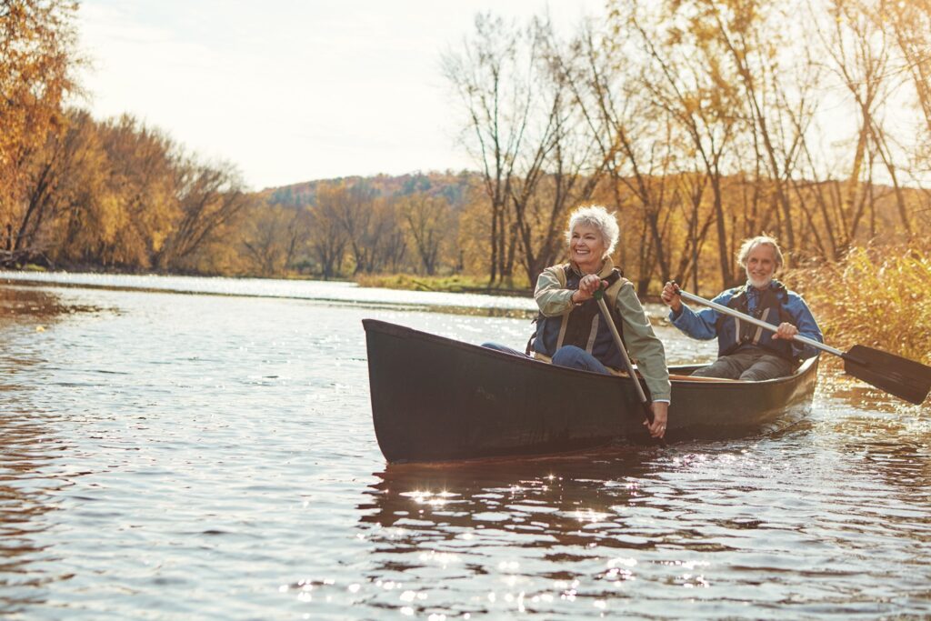 An older couple paddles a canoe together on a calm river surrounded by autumn trees. Both are smiling and wearing life jackets as sunlight reflects off the water.