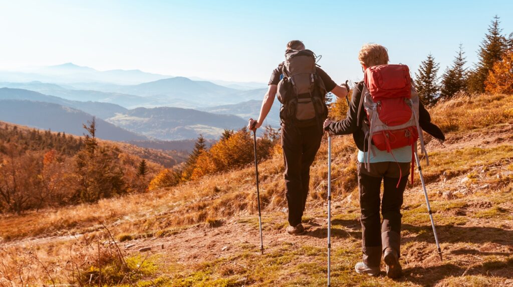 Two hikers with backpacks and trekking poles walk up a mountain trail surrounded by autumn-colored trees. The landscape stretches into distant rolling hills and misty blue mountains under a clear sky.