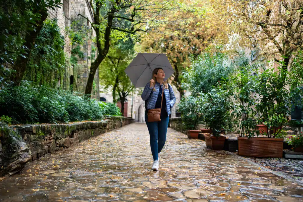 A woman walks alone along a charming, rain-soaked cobblestone path lined with greenery and stone walls, holding a grey umbrella. She wears a blue puffer jacket and looks upward with a calm expression, surrounded by trees with autumn leaves in a peaceful, historic setting.