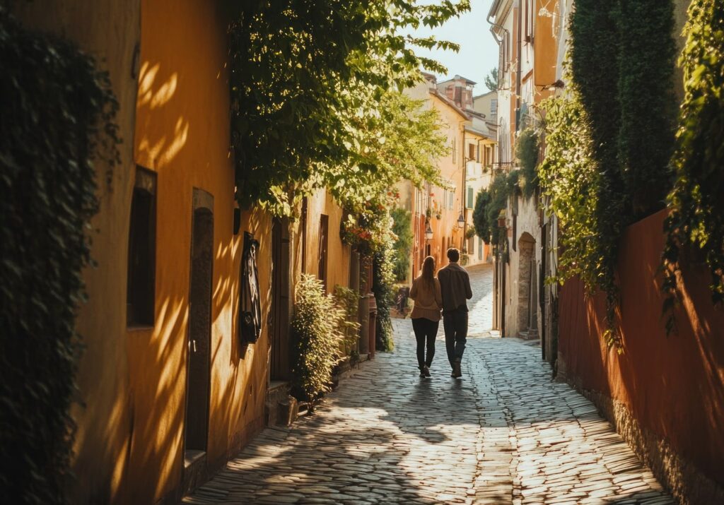 A couple walks hand in hand down a charming, narrow cobblestone street lined with colorful buildings and lush greenery. Warm sunlight filters through the trees, casting soft shadows and highlighting the peaceful, romantic atmosphere of the quiet European alleyway.