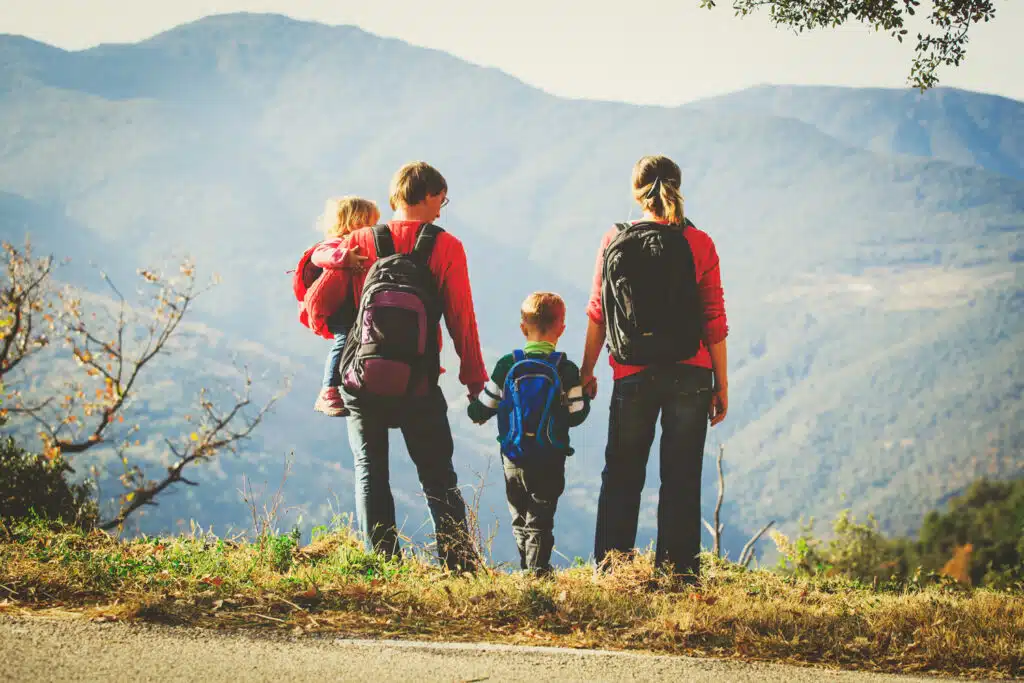 A family of four stands at the edge of a scenic overlook, facing a vast mountain landscape. The parents, both wearing backpacks, hold hands with their two young children, one of whom is being carried, as they take in the view together.