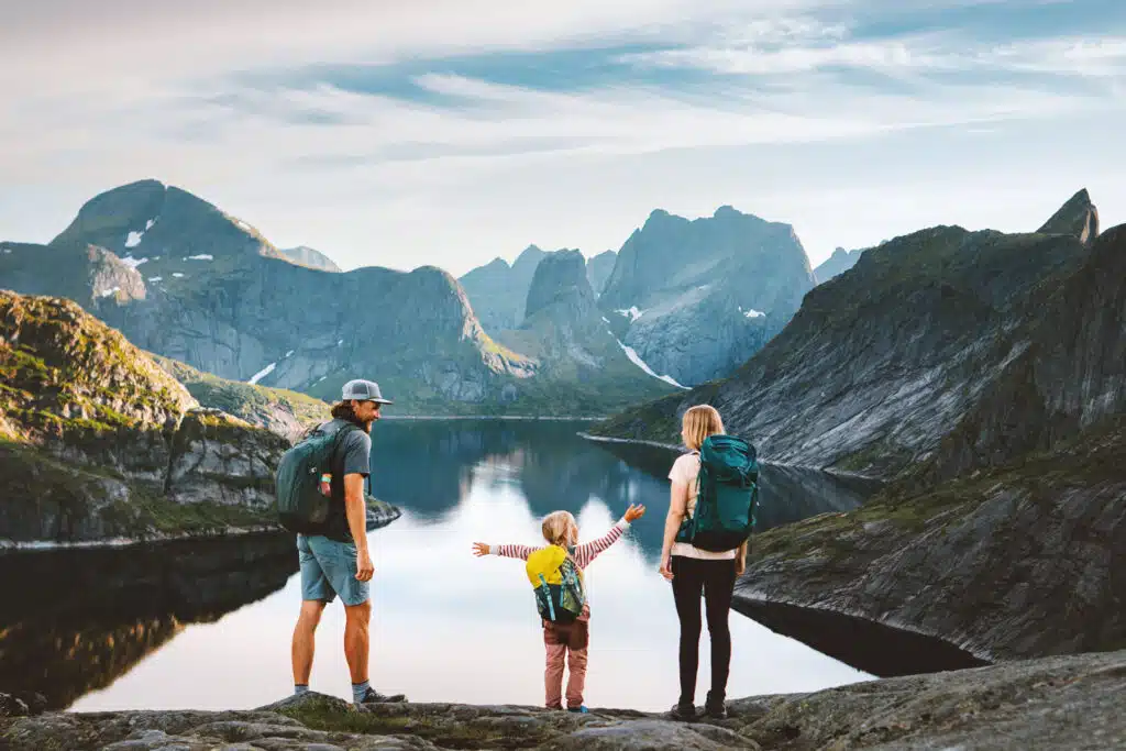 A family of three stands on a rocky ledge overlooking a serene mountain lake surrounded by towering peaks, with the child joyfully stretching out their arms. All are wearing backpacks, suggesting they’ve hiked to this scenic viewpoint under a bright, lightly clouded sky.