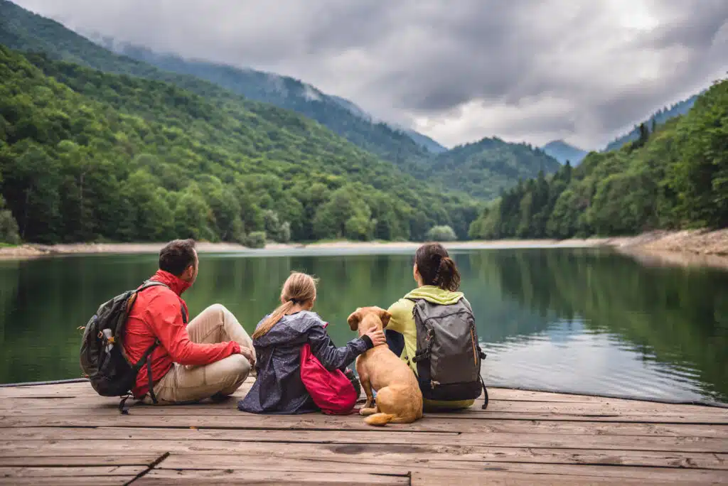 A family sits on a wooden dock overlooking a calm lake surrounded by lush green mountains and a cloudy sky. A child gently pets their dog as all three, wearing backpacks and outdoor gear, take in the peaceful natural scenery.