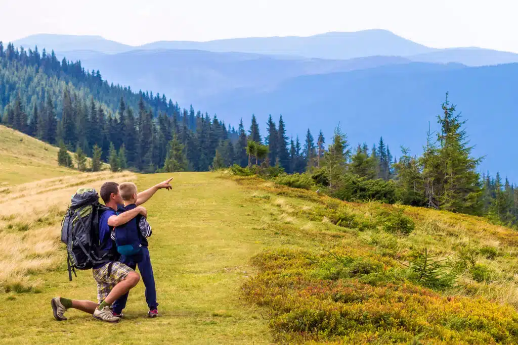 A man and child pause on a grassy mountain trail surrounded by evergreens, with the man kneeling and pointing toward the distant blue ridges. Both wear backpacks, suggesting they’re on a hiking adventure through a scenic, high-altitude forest.