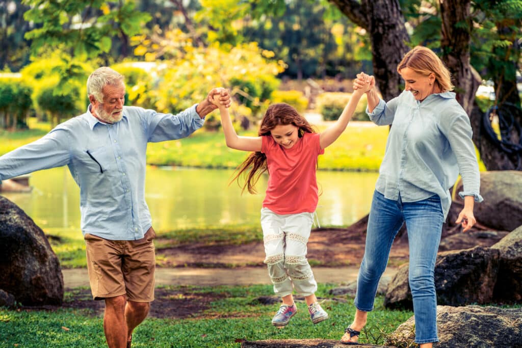 A joyful scene in a park shows an older man and a woman swinging a young girl between them as they walk. The child is mid-air, smiling with delight, while the adults beam with happiness. Behind them, a serene pond and lush greenery create a peaceful, sunlit backdrop.