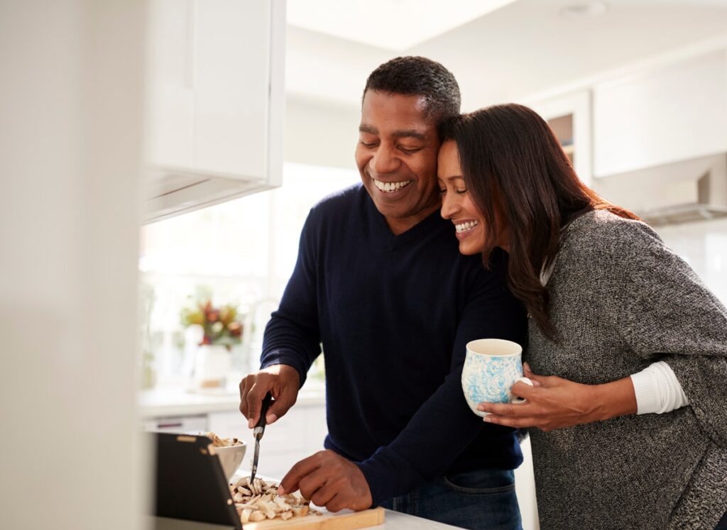 A couple smiles warmly in a bright kitchen as the man chops mushrooms on a cutting board while the woman leans in close, holding a patterned mug. They appear to be enjoying a relaxed moment together, cooking with a tablet propped up nearby.