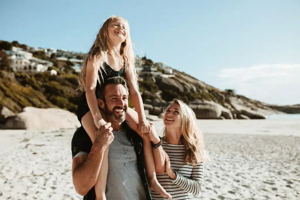 A happy family enjoys a sunny day at the beach, with a man carrying a smiling young girl on his shoulders and a woman standing beside them looking up joyfully. Behind them, sandy hills and coastal homes create a relaxed, scenic backdrop under a clear blue sky.