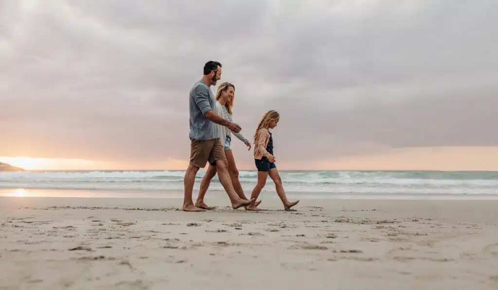 A family of three walks barefoot along the shoreline at sunset, with gentle waves rolling in behind them and a soft, pastel sky above. The child leads the way, followed by her smiling parents, all enjoying a peaceful moment on the beach.